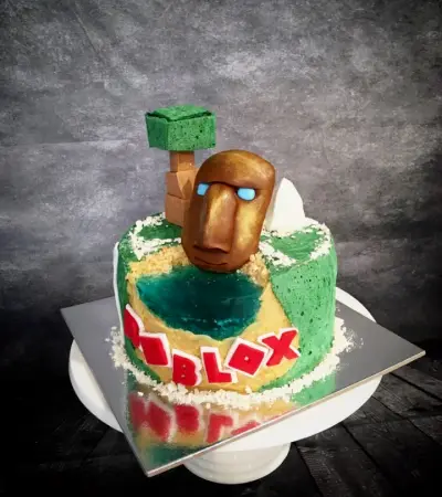 Cylindrical cake with green and gold sides, a teal 'lake', a bronze blocky face in sunglasses, a green block tree, and red 'ROBLOX' letters.