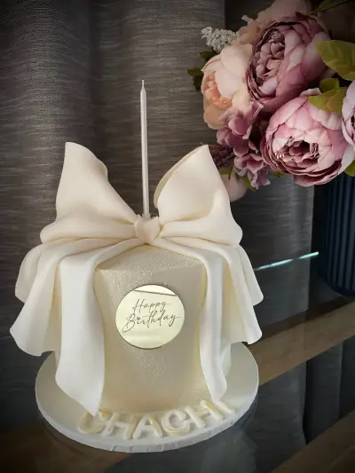 Ivory cake styled as a wrapped gift with a large fondant bow, a gold Happy Birthday plaque, and Chachi on the base board.