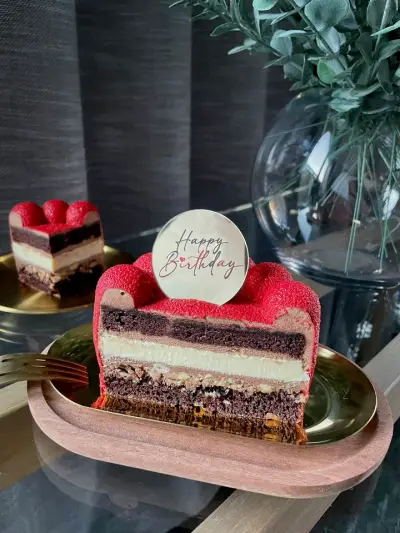 A slice of cake with layers of chocolate sponge, cream and crumbly biscuit under a glossy red domed top and a Happy Birthday topper.
