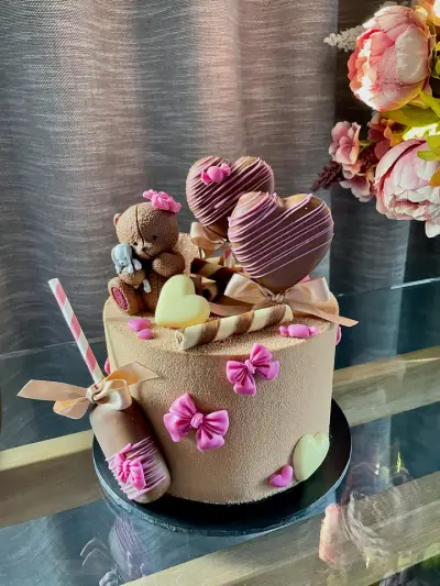 A brown cake decorated with pink fondant bows, a teddy bear figurine holding a toy, chocolate-dipped heart lollipops and a wrapped treat.