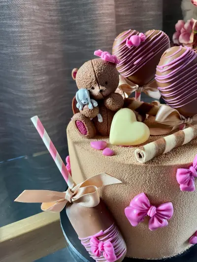 Close-up of a teddy bear figurine with a toy, a pale heart, chocolate-dipped hearts with pink drizzle and pink bows on a brown cake.