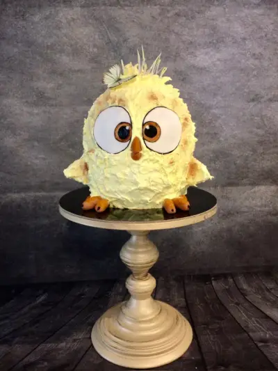 Round cake shaped like a fluffy yellow chick with oversized cartoon eyes, an orange beak, wings and feet, and a small butterfly on top.
