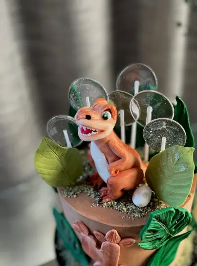 A fondant baby dinosaur sits among green leaves, a white egg and sparkly disc lollipops on top of a brown cake.