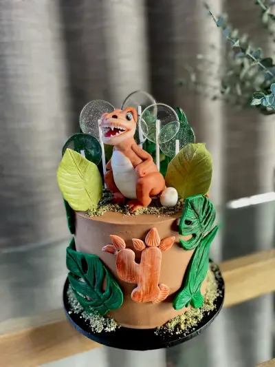 A brown cake with a textured orange number 4, topped with a fondant dinosaur, leaves, an egg and sparkly disc lollipops.