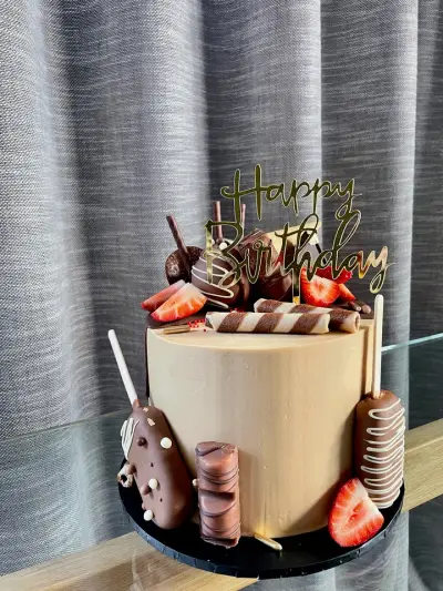 A smooth tan cake with a gold Happy Birthday topper, chocolate-dipped strawberries, a wafer roll and chocolate-coated cake pops.