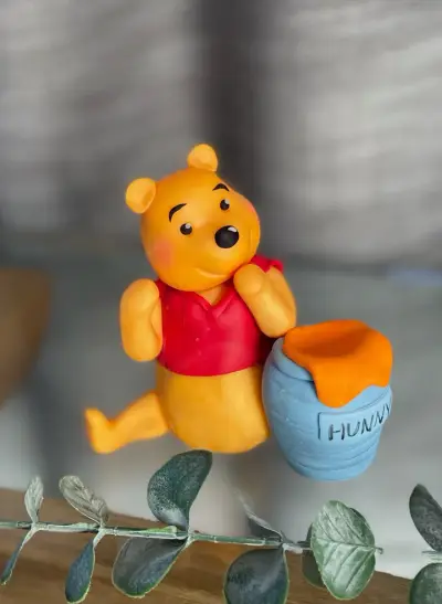 A fondant bear figurine in a red top reaches toward a honey pot labelled 'Hunny', displayed among greenery.