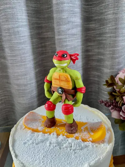 A fondant turtle superhero figure with a red mask, shell guard and 'R' emblem stands on a snowboard atop a cake.
