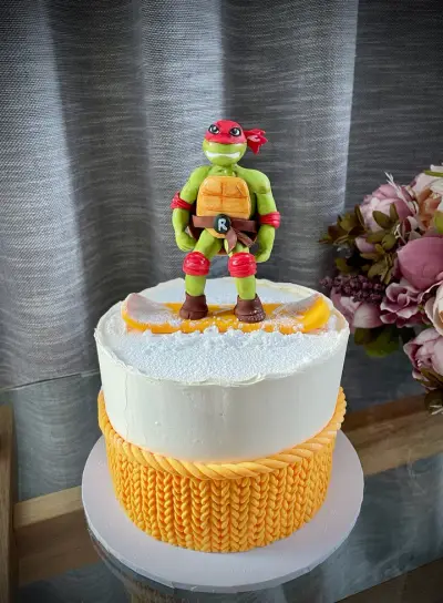 A two-tier cake with a snow-textured white top and an orange knitted-pattern base, topped with a turtle superhero on a snowboard.