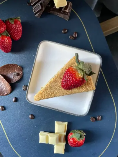 A triangular slice of crumbed sponge cake topped with a strawberry, plated with fresh strawberries, chocolate and coffee beans.