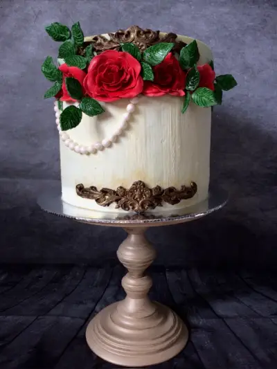 Round cake with rustic cream buttercream, a cluster of red sugar roses and green leaves on top, a pearl strand, and bronze baroque trim.