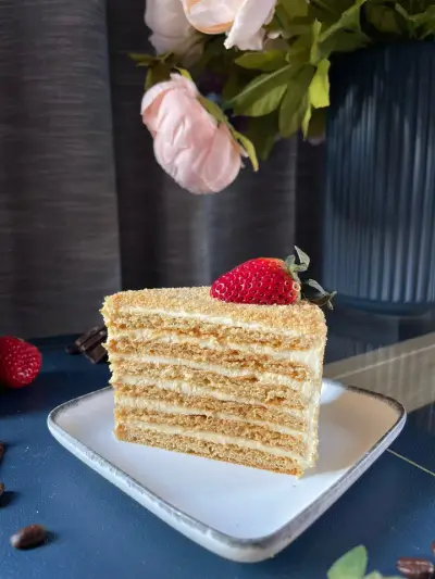 A slice of cake with many thin golden layers and cream filling, topped with a fresh strawberry, served on a white plate.