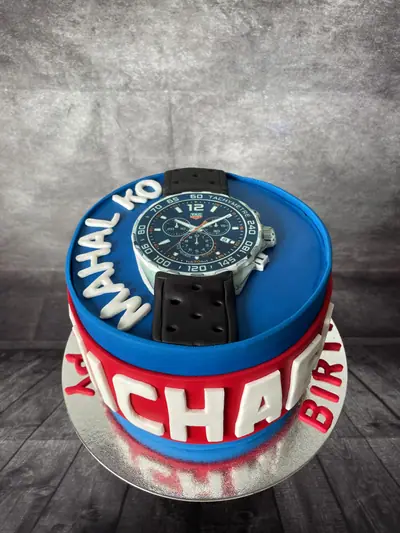 Round red-and-blue cake topped with a sculpted black-strap TAG Heuer chronograph watch, with large white lettering around the sides.