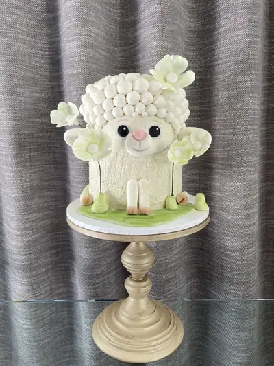 Sculpted white lamb cake with round wool curls, black eyes, a pink nose, front legs, pale-green sugar flowers and a green grass-effect base.