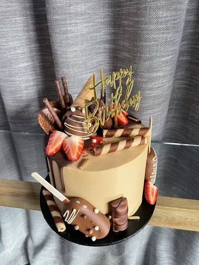 Caramel-coloured birthday cake with dark chocolate drip, strawberries, chocolate-coated cakesicles, wafer rolls, chocolate discs, chocolate pieces, red sprinkles and a gold topper.