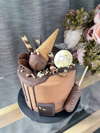 Milk chocolate cake with dark chocolate drip, an upside-down ice cream cone over a chocolate-coated dome, wafer rolls, chocolate discs, a cakesicle, chocolate bar and gold pearls.