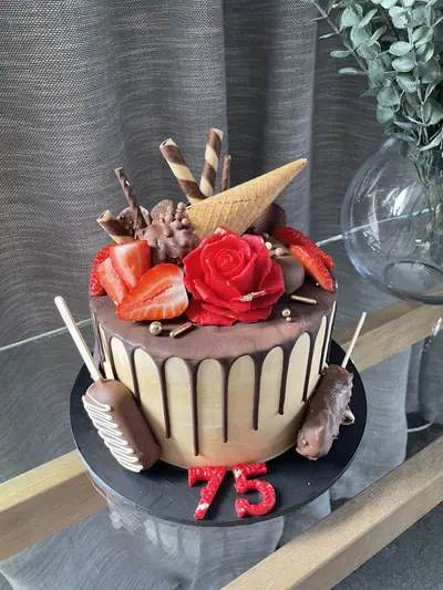 Ivory 75th birthday cake with dark chocolate drip, a large red rose, strawberries, chocolate-coated cakesicles, wafer rolls, an ice cream cone, gold pearls and a red number 75.