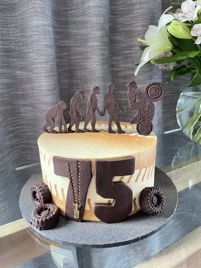 75th birthday cake with chocolate silhouettes showing human evolution ending in a motorbike wheelie, a large chocolate number 75 with zipper detail, and chocolate tyre decorations.