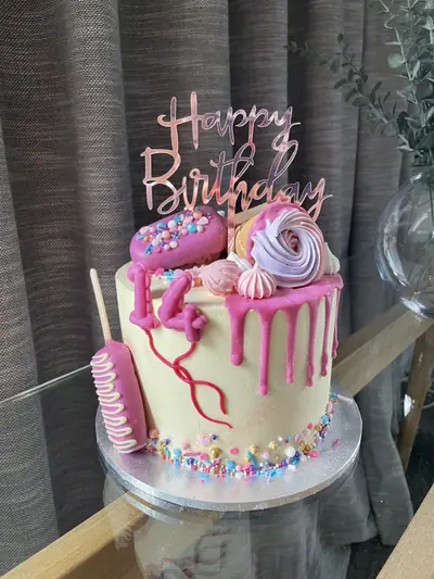Tall white 14th birthday cake with hot-pink drip, pink number candles, a pink cakesicle, pink and lilac meringues, multicoloured sprinkles and a rose-gold birthday topper.