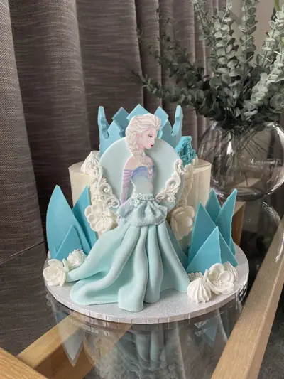 Frozen-inspired white cake with a sculpted Elsa figure in a flowing pale-blue gown, turquoise ice shards, white sugar flowers and ornate white scrollwork.