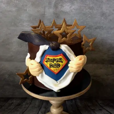 Round chocolate-brown cake with a torn-shirt design revealing a 'SUPER DAD' shield, hands, a cape, and gold stars around the top.