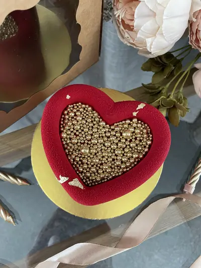 Top view of a red heart-shaped velvet-textured cake with a recessed heart centre filled with metallic gold sugar pearls and decorated with gold leaf.
