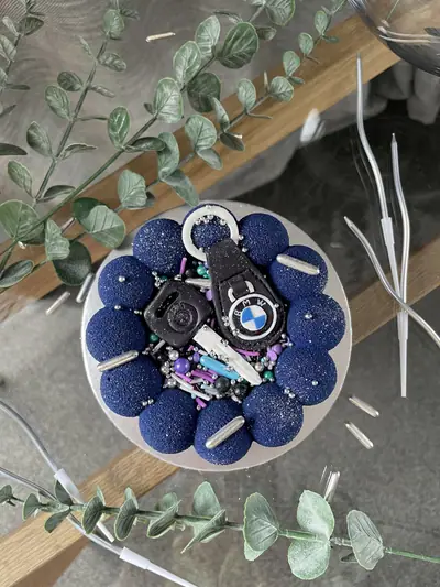 Top view of a round cake edged with dark navy textured spheres and topped with edible BMW car key fobs, a silver ring and key blade, colourful sprinkles and metallic sugar pearls.