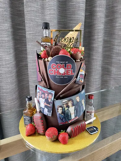Two-tier chocolate birthday cake with Cold Chisel album images, a gold birthday topper, miniature whisky and vodka bottles, strawberries, chocolate treats, an ice cream cone and music-themed decorations.