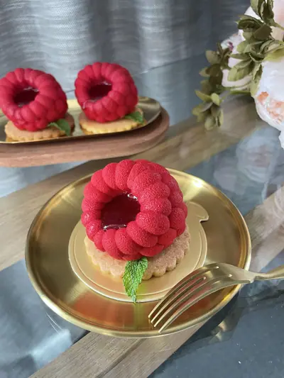 Three individual raspberry-shaped red desserts with dimpled velvet coating and glossy raspberry centres, each on a fluted golden tart with a mint leaf.
