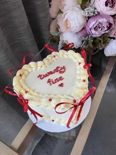 White heart-shaped cake with vintage piped borders, red ribbon bows, red sugar pearls and red script reading ‘twenty fine’.