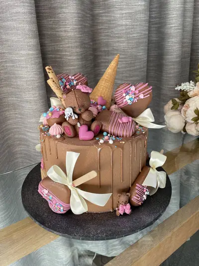 Chocolate drip cake topped with a sculpted brown teddy bear holding a white bunny, an ice cream cone, chocolate-coated cakesicles, meringues, bows and pink-and-blue sprinkles.