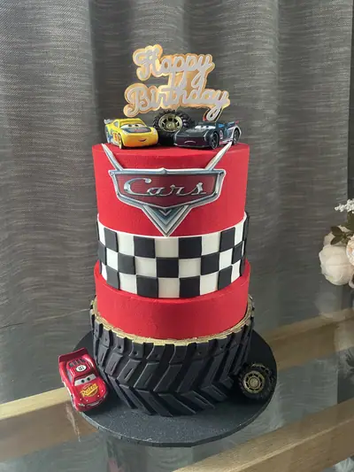 Three-tier red Cars-themed birthday cake with a black tyre-tread base, black-and-white chequered middle tier, Cars logo, racer character toppers, loose tyres and a gold birthday topper.