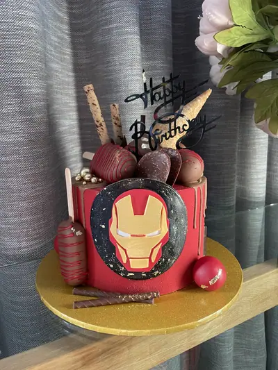 Red Iron Man-themed cake with a gold-and-red helmet emblem, black birthday topper, red chocolate drip, ice cream cone, chocolate discs, cakesicles, wafer rolls and gold leaf.