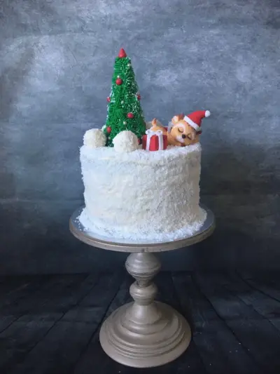 Round cake coated in white coconut-look icing, topped with a Christmas tree, a red gift box, snowballs, and a sleeping fawn in a Santa hat.