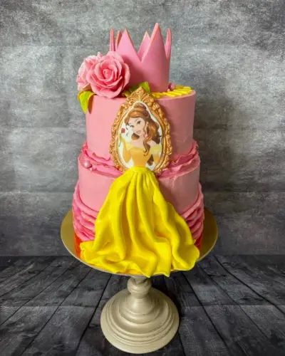 A two-tier pink cake with a fondant crown, pink rose, and a Belle image above a flowing yellow fondant skirt.