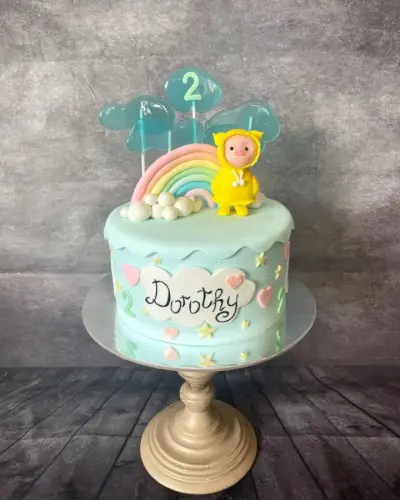 Mint cake with a piped name and a number 2, a pastel rainbow, clouds, hearts, stars, and a raincoat-wearing piglet figure.