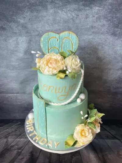 Two-tier mint cake with a piped name and 'Birthday', a wood-effect '60' heart topper, cream roses, and white rope trim.