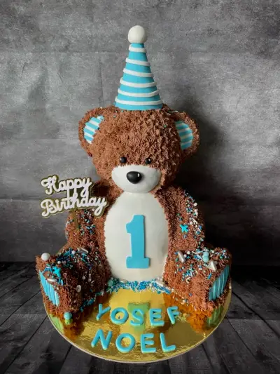 Cake shaped like a brown teddy bear in a striped party hat, with a blue number 1 and a piped name on the gold board.