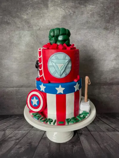 Two-tier cake with an Iron Man reactor and Hulk fist on top, a Captain America shield, star-striped tier, and Thor's hammer.