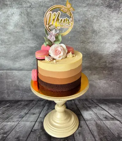Ombre brown cake topped with pink macarons, a pale pink rose, meringue kisses, and a gold 'Mum' topper with a butterfly.