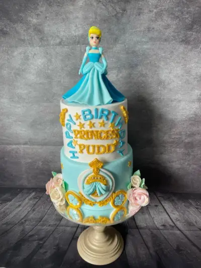 Two-tier cake with a Cinderella figure, 'Happy Birthday Princess Pudd' lettering, a gold crest, and pink and white roses.