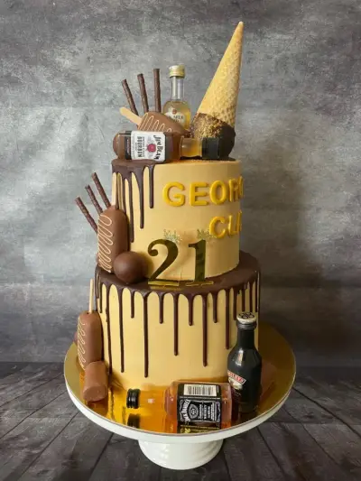 Two-tier caramel drip cake decorated with miniature spirit bottles, chocolate sticks, an ice cream cone, and a gold '21'.