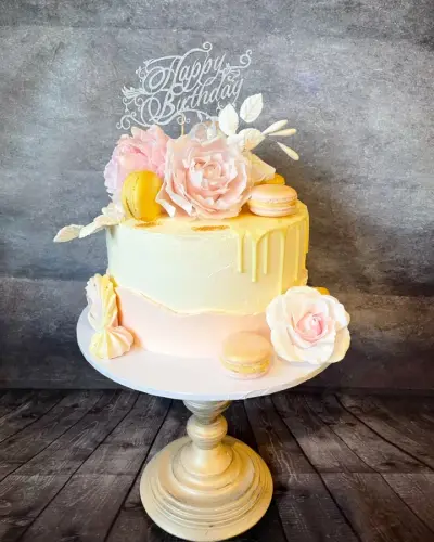 Single-tier cream cake with a white chocolate drip, pink sugar roses, yellow macarons and a Happy Birthday topper.