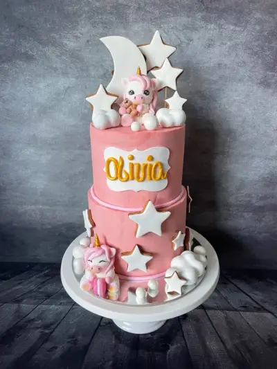 Two-tier pink cake named Olivia, decorated with a unicorn figure, white stars, a crescent moon and fondant clouds.