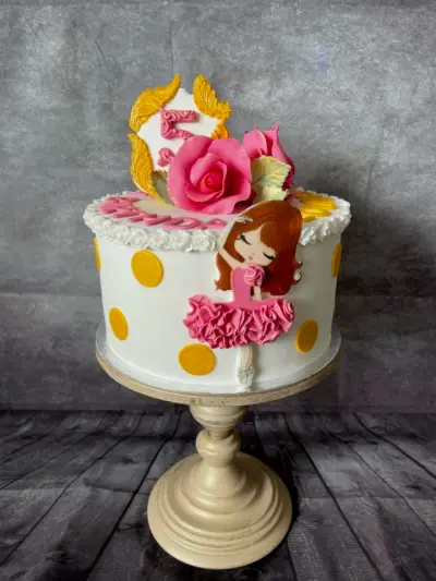 White cake with gold polka dots, pink sugar roses, a gold number 5 topper and a cartoon ballerina figure.