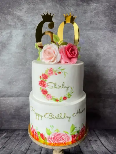 Two-tier white cake with a gold 60 topper, pink roses, and Happy Birthday Mama and Shirley written in icing.