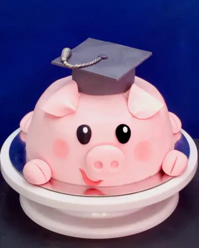 Dome-shaped cake styled as a pink pig's face with black eyes, blushed cheeks, a snout, ears, and a grey graduation cap with tassel.