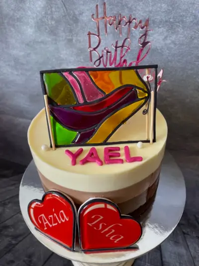 Cream and tan cake with a stained-glass style topper, a Happy Birthday sign, the name Yael, and heart cookies reading Azia and Isha.