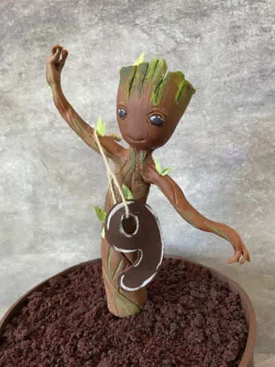 Close-up of a Baby Groot figure holding a number 9 tag, standing in chocolate cookie-crumb soil on top of a cake.