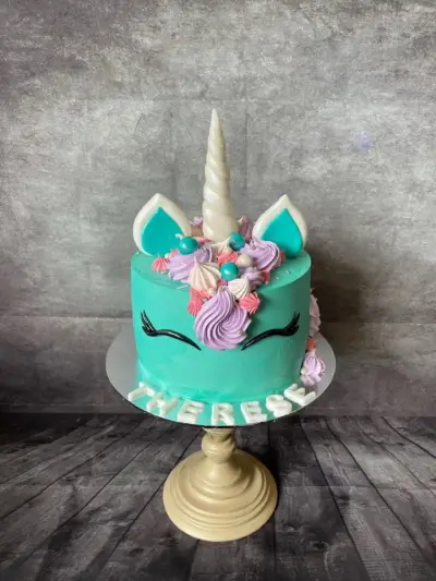 Turquoise unicorn cake with a white horn, pastel purple and pink piped swirls, lash eyes, and a name in white letters.