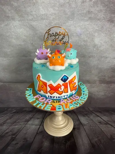 Teal cake with monster figures on clouds, an AXIE INFINITY logo, a gold happy birthday topper, and colourful lettering at the base.